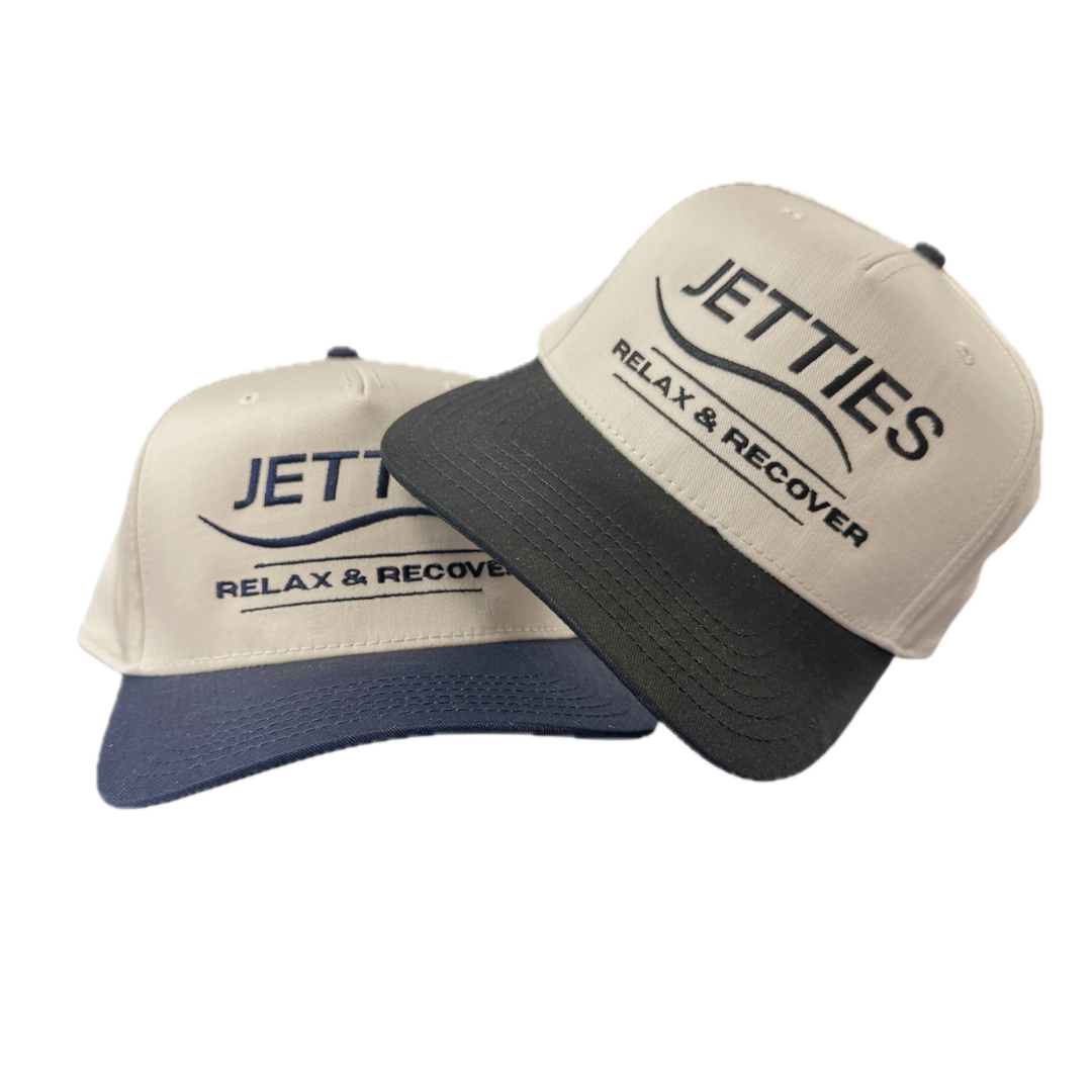 Two JETTIES Relax & Recover snapback hats in tan with navy and black brims — embroidered front logos, side-by-side product display.