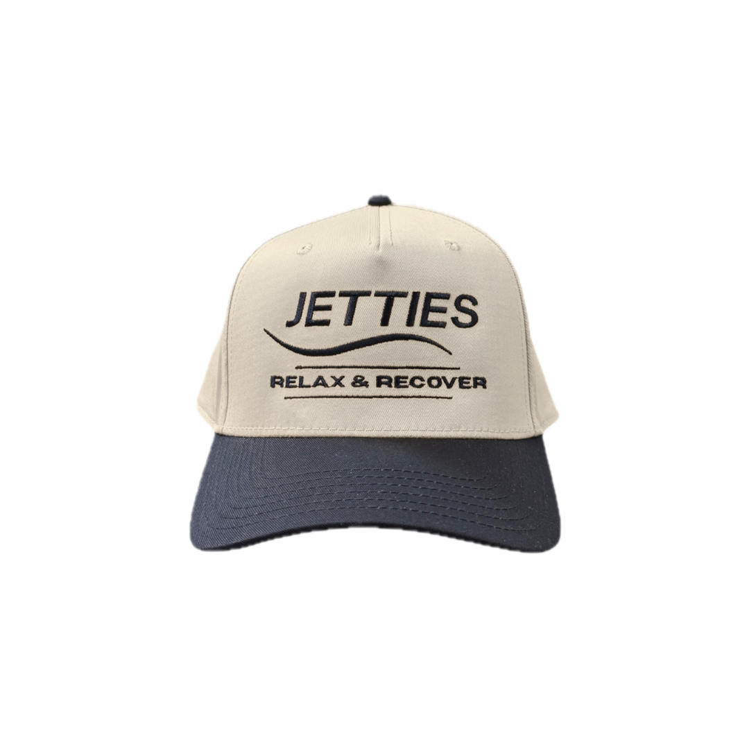 JETTIES Relax & Recover snapback hat with navy brim — structured beige crown and embroidered logo, front view.