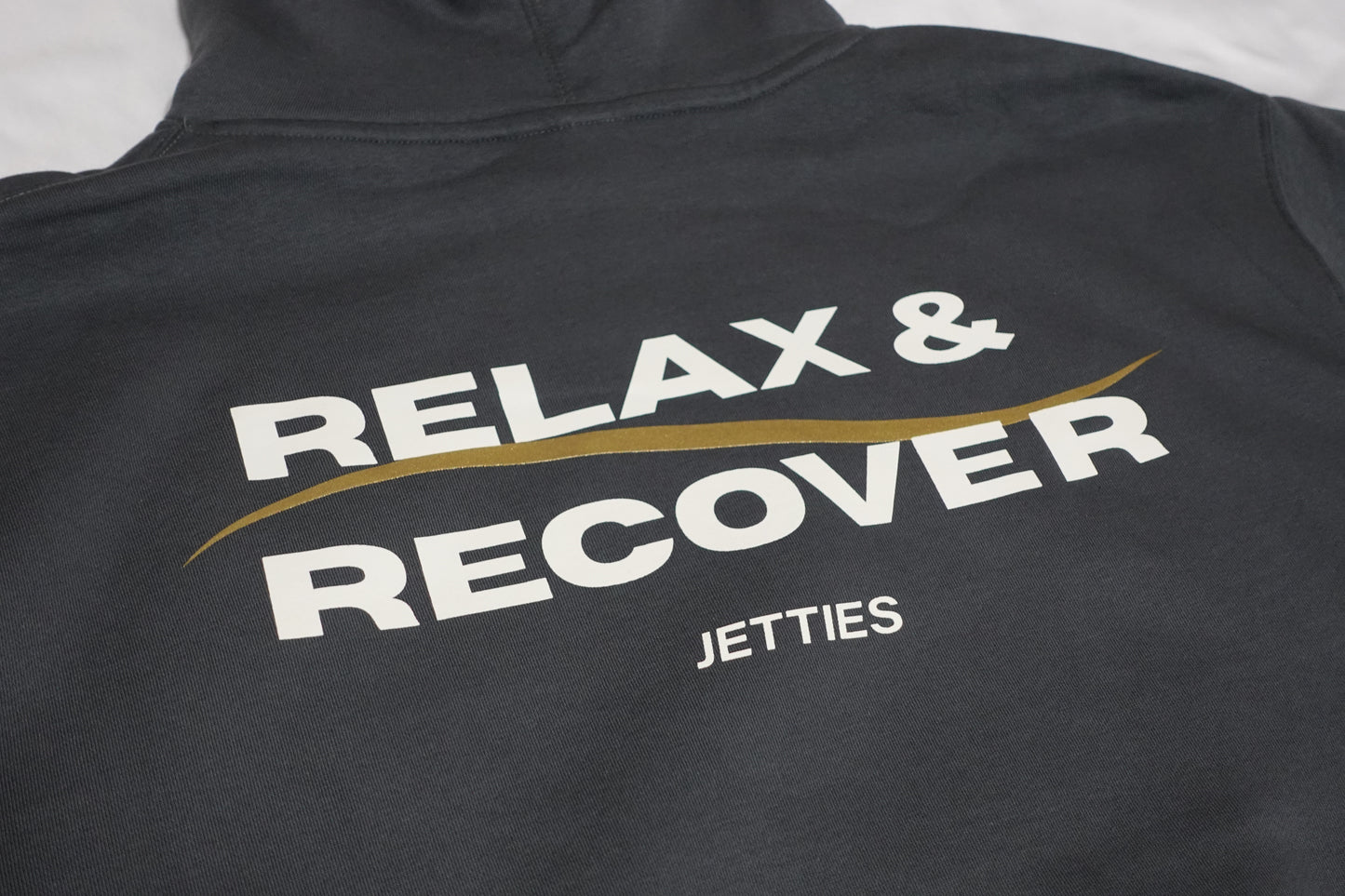 Close-up angled shot of the Relax & Recover back graphic showing the white lettering and gold wave detail on a gray hoodie.