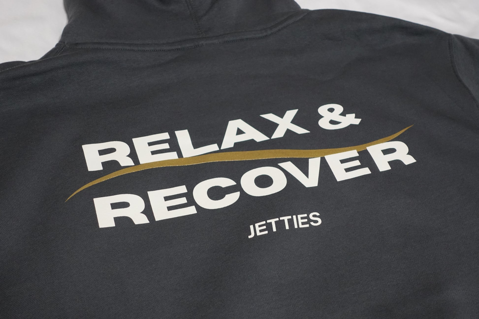 Close-up angled shot of the Relax & Recover back graphic showing the white lettering and gold wave detail on a gray hoodie.