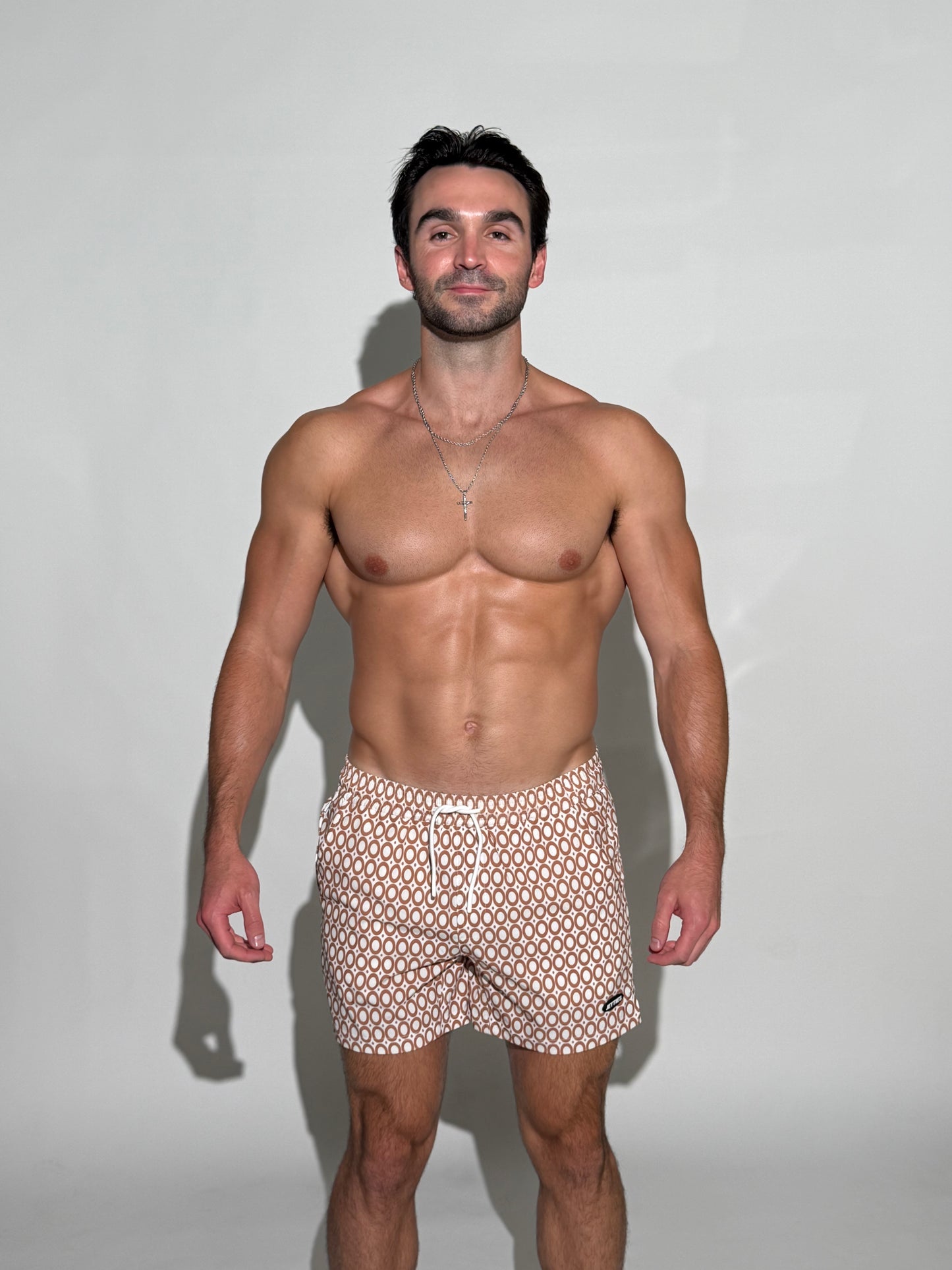 Model wearing the JETTIES Echo Caramel, brown and white circles mens bathing suit, geometric pattern