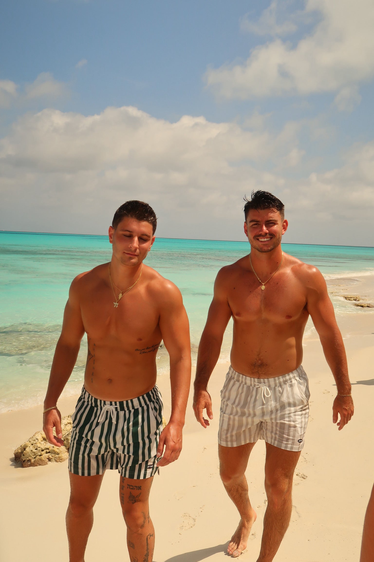 Two men standing on a beach with turquoise water and blue sky wearing JETTIES CODE GREEN & CODE GRAY PLUNGE SHORT BATHING SUITS
