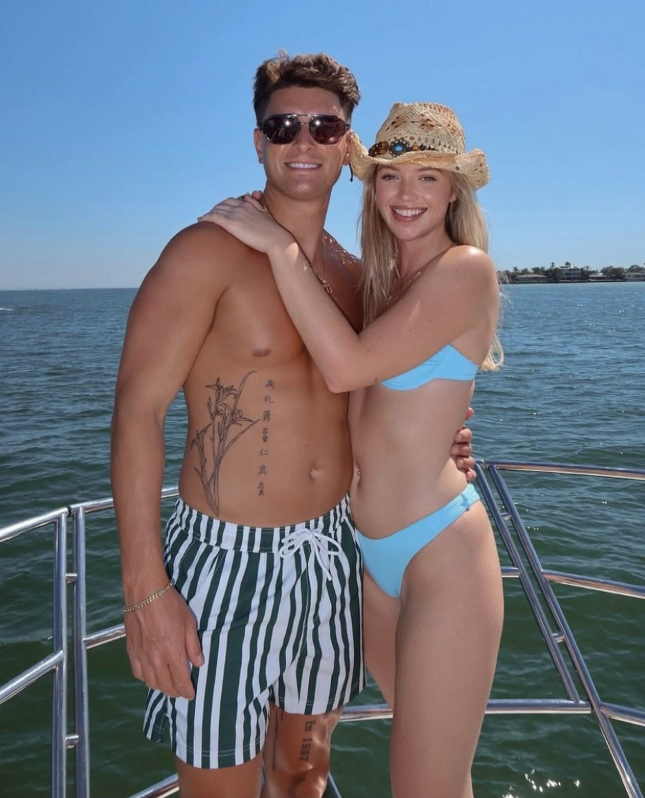 Couple standing out on edge of boat with man wearing JETTIES CODE GREEN PLUNGE SHORT bathing suit