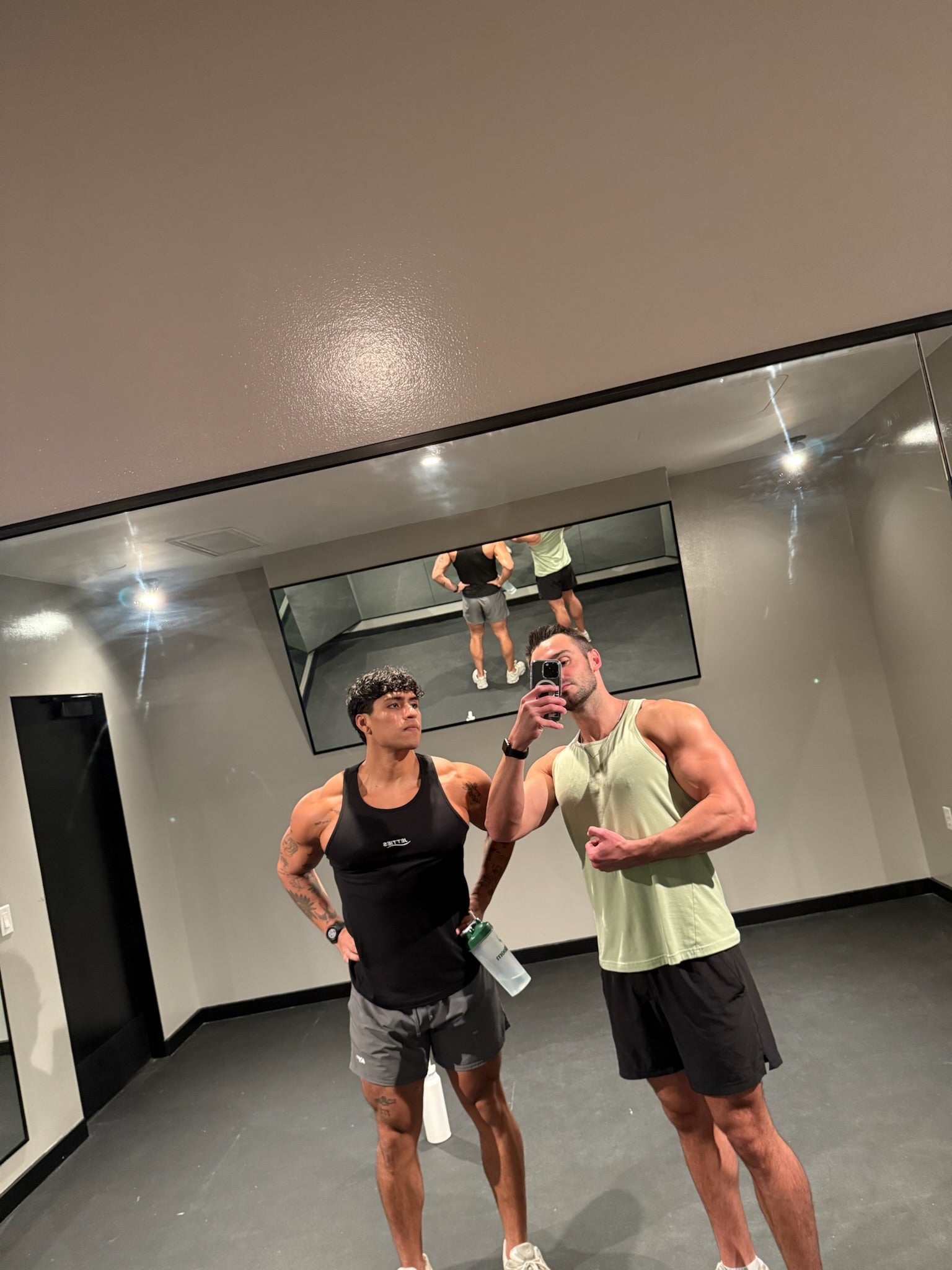 Two muscular individuals taking a selfie in a mirror rocking JETTIES ASCEND TANK TOPS BLACK & MATCHA GREEN.