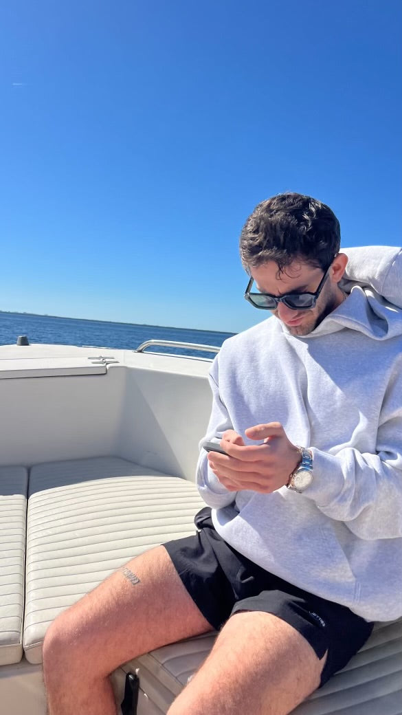 Man sitting on a boat using a phone with clear blue sky and water in the background wearing JETTIES CLASSIC BLACK PLUNGE SHORT SWIMSUIT