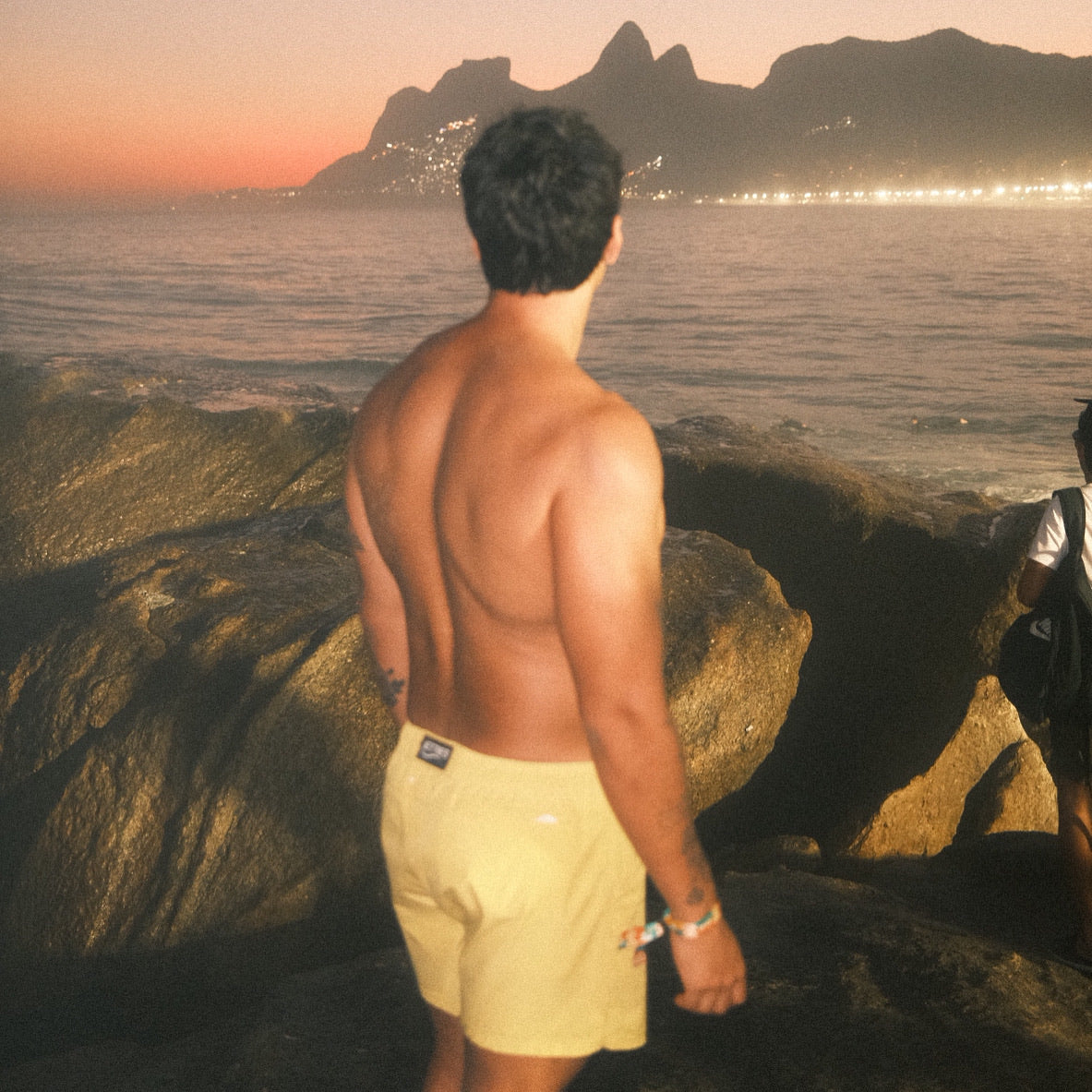 Man standing on a rocky outcrop overlooking a scenic landscape with mountains and water at sunset wearing JETTIES SUNSET YELLOW PLUNGE SHORT swimwear