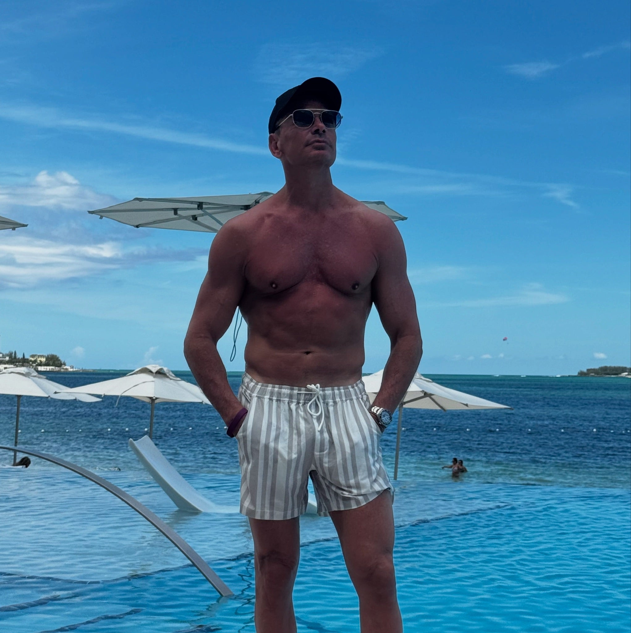 Man standing by a pool with clear blue water and a blue sky wearing JETTIES CODE GRAY PLUNGE SHORT bathing suit