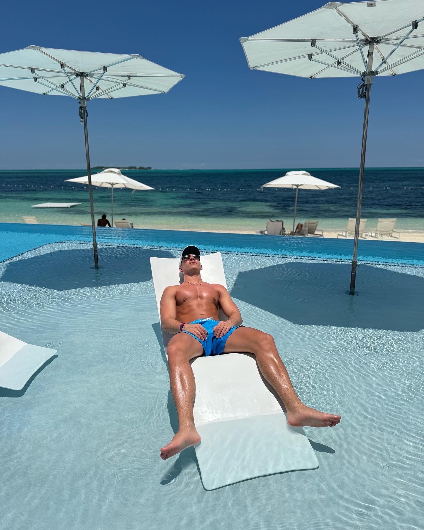 Man lounging on a pool float by a pool with ocean view tanning wearing JETTIES BLUE PLUNGE SHORT bathing suit