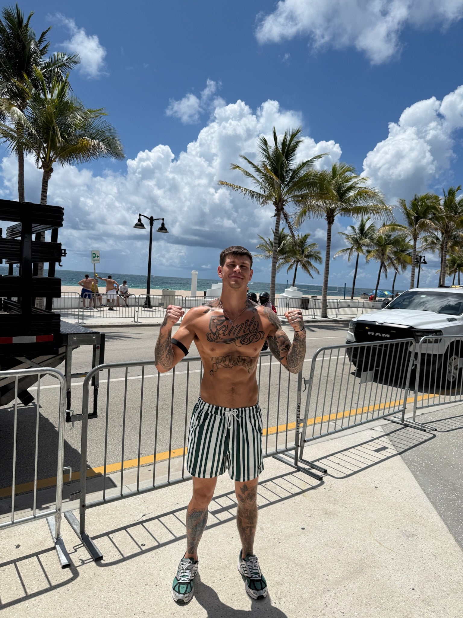 Man flexing muscles on a street with palm trees and ocean in the background rocking JETTIES CODE GREEN PLUNGE SHORT swim suit