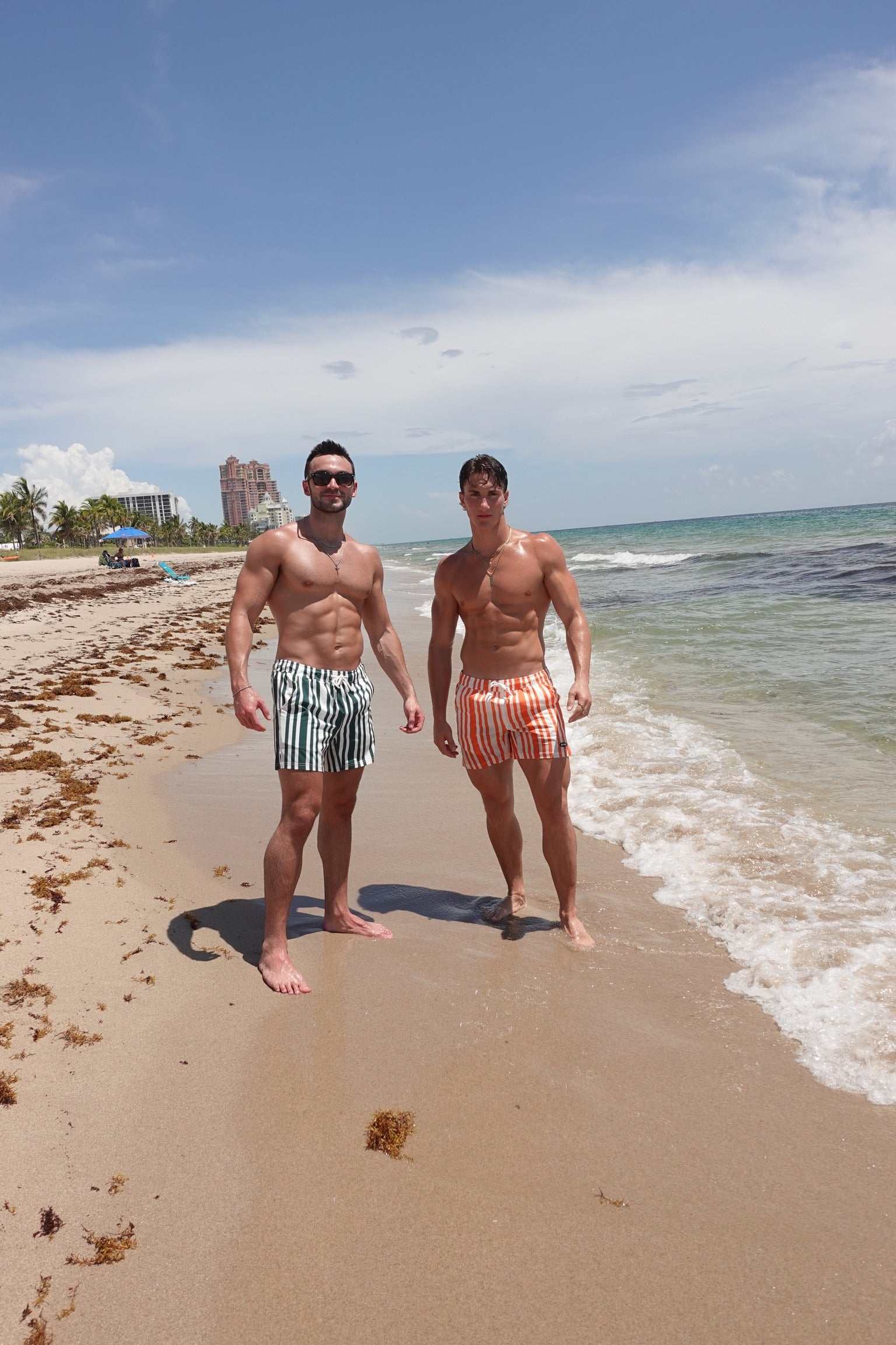 Two men standing on a beach with ocean waves and clear sky wearing JETTIES CODE GREEN & CODE ORANGE PLUNGE SHORTS striped swimwear